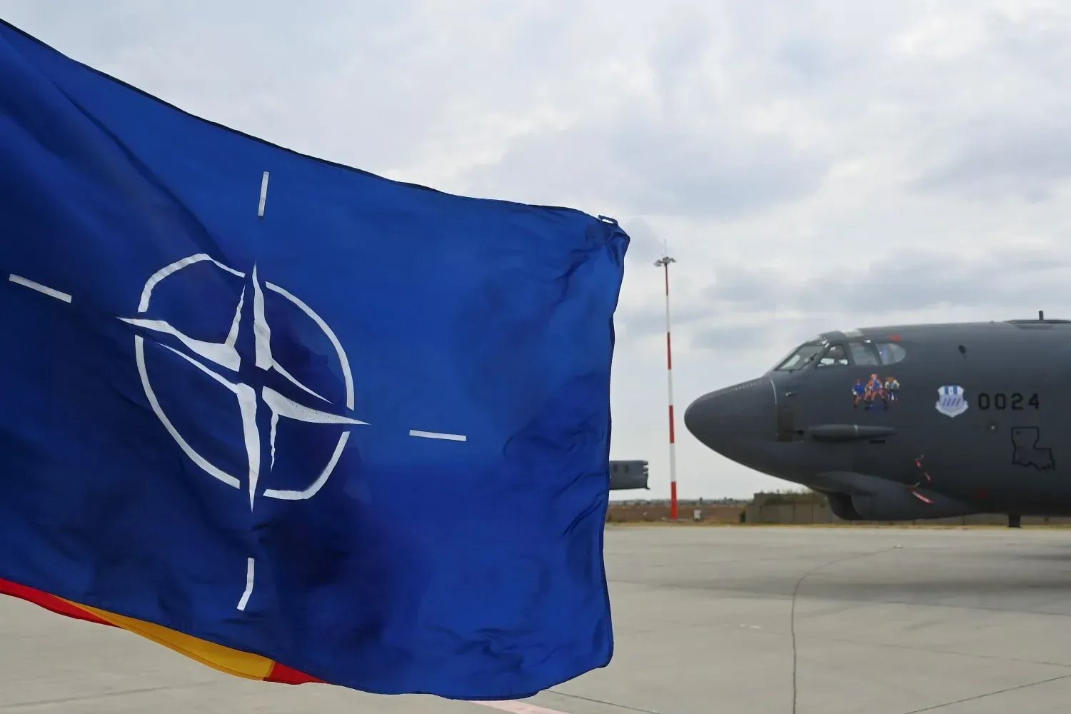 Steag NATO fluturând lângă un avion militar pe o pistă de aeroport.