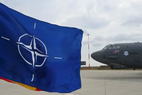 Steag NATO fluturând lângă un avion militar pe o pistă de aeroport.