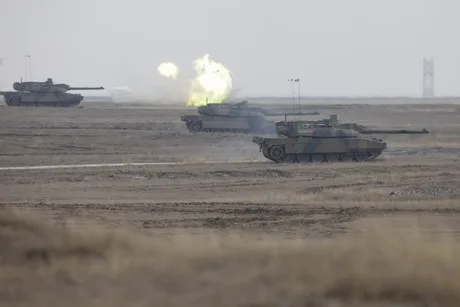 Tancuri în exerciții militare pe un teren deschis, fum și foc în fundal.
