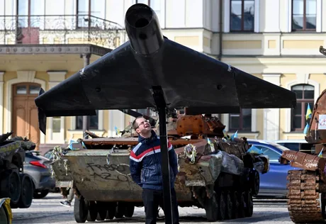 Un bărbat observă o dronă expusă lângă vehicule militare vechi.
