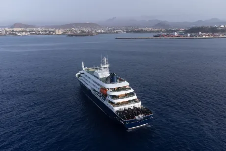 Nava MV Hondius navigând pe ape, cu orașul în fundal.