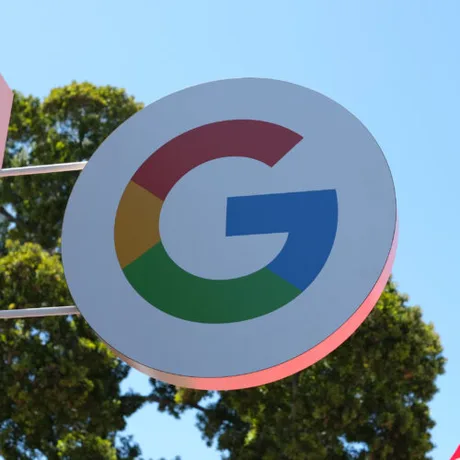 Logo-ul Google expus într-un peisaj urban, cu copaci verzi în fundal.