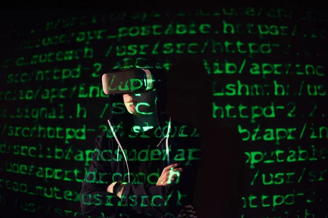 Un hacker cu ochelari VR stând în fața unui ecran plin de cod.