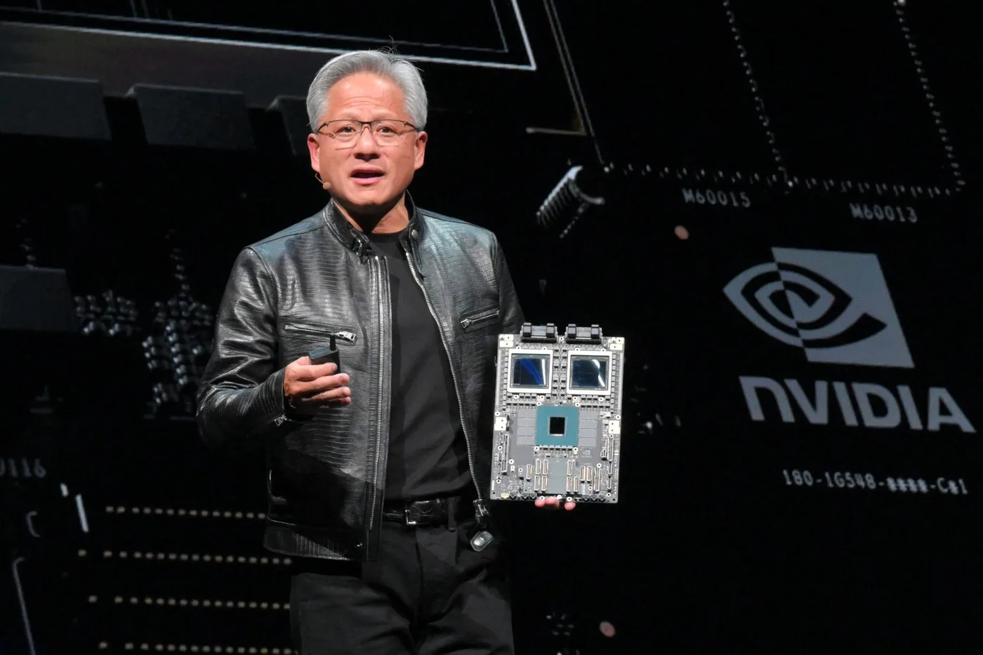 Jensen Huang prezentând o placă de bază pe scenă, purtând o geacă de piele neagră.