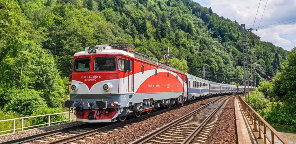 Tren CFR circulând pe o linie ferată înconjurată de natură verde.