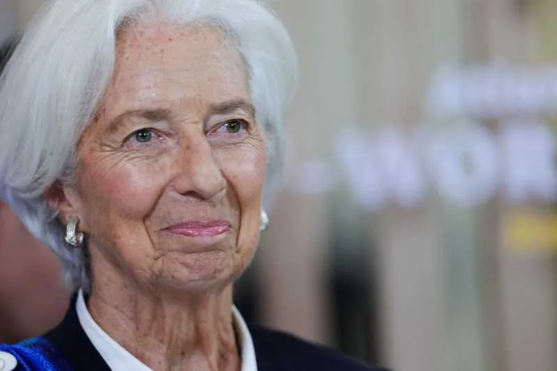 Christine Lagarde zâmbind, într-un cadru oficial, cu părul alb și o vestă elegantă.