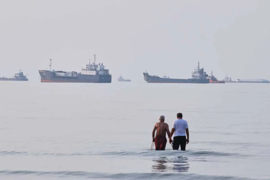 Două persoane merg pe plajă, în fundal se văd nave pe apă.