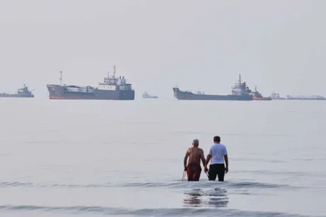 Două persoane merg pe plajă, în fundal se văd nave pe apă.