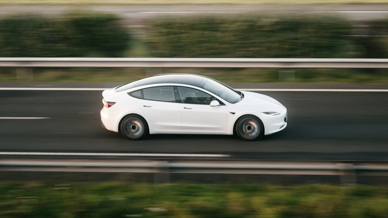 Sedan electric Tesla Model 3 în mișcare pe autostradă, cu peisaj verde în fundal.