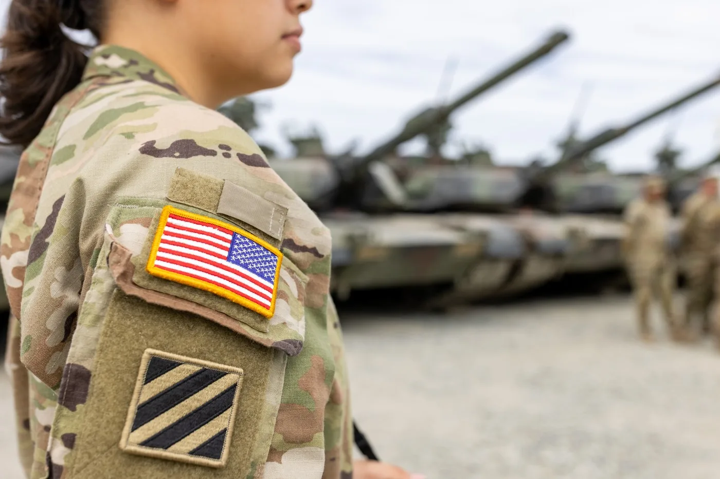 Soldat american în uniformă, cu tancuri în fundal, pregătiri militare în desfășurare.