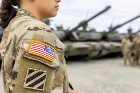 Soldat american în uniformă, cu tancuri în fundal, pregătiri militare în desfășurare.