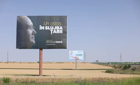 Panou publicitar cu Nicolae Ciucă, pe fundalul unui peisaj rural.