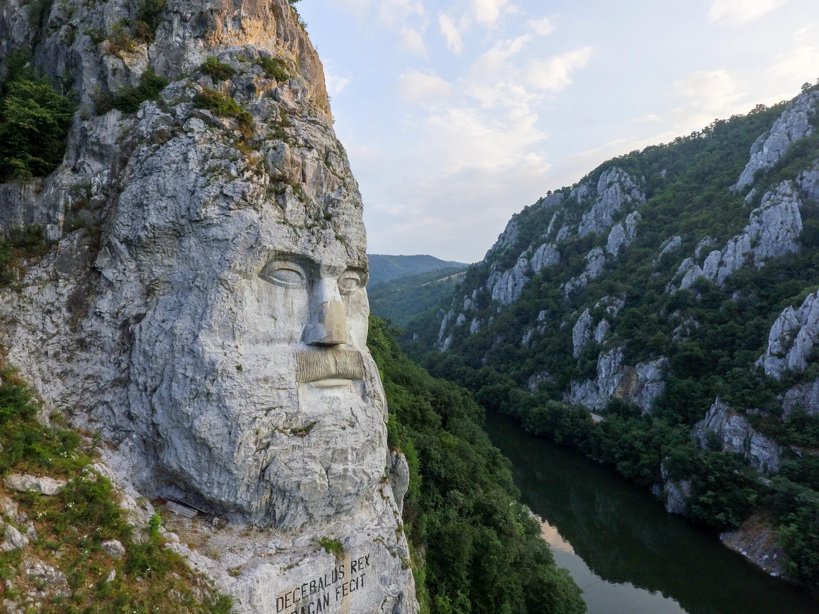 Statuia lui Decebal sculptată în stâncă, pe malul Dunării, înconjurată de peisaje montane verzi.