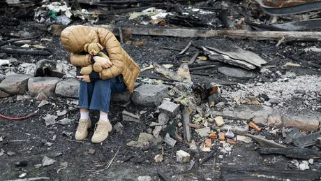 Un copil îmbrăcat călduros stă pe ruine, ținând un ursuleț de pluș.