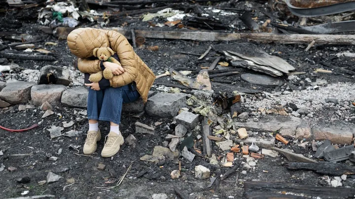 Un copil îmbrăcat călduros stă pe ruine, ținând un ursuleț de pluș.