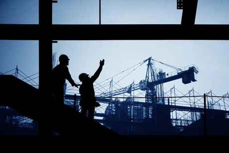 Lucrători pe un șantier de construcții, cu schele și macarale în fundal.