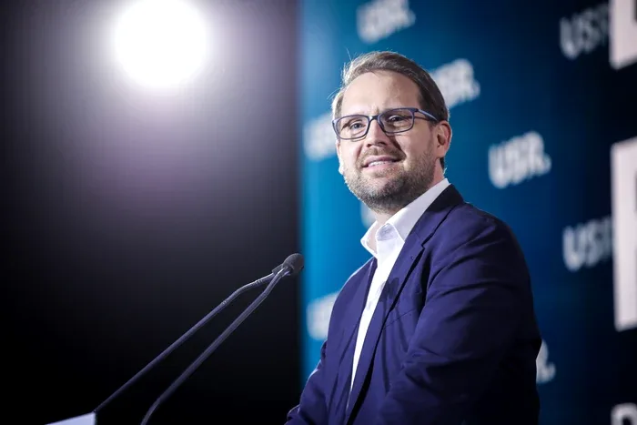 Dominic Fritz vorbește la o conferință, cu steaguri în fundal.