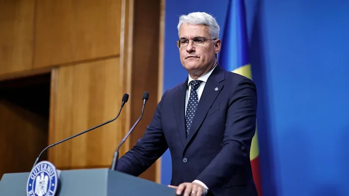 Dragoș Pîslaru susține un discurs în fața presei, cu steagul României în fundal.