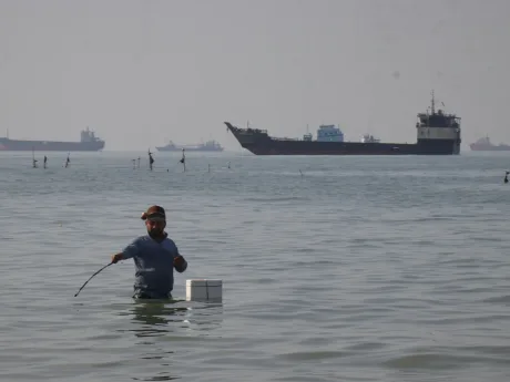 Un bărbat pescuiește în apă, în fundal se văd nave cargo.