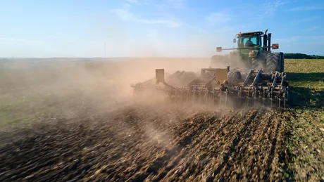 Tractor lucrând pe câmp în condiții de praf și soare.