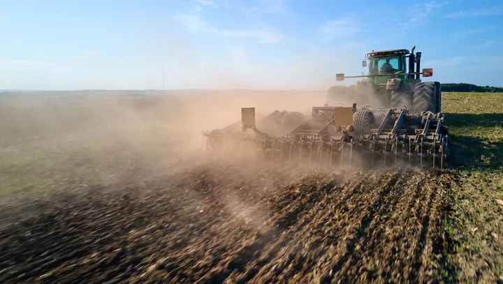 Tractor lucrând pe câmp în condiții de praf și soare.