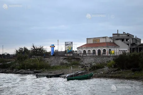 Peisaj de coastă cu bărci și clădiri în fundal, sub un cer înnorat.