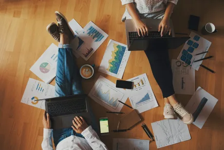 Persoane lucrând pe laptopuri, înconjurate de grafice și documente financiare.
