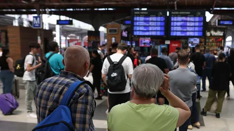 Oameni așteptând în Gara de Nord din București, cu panouri de informații în fundal.