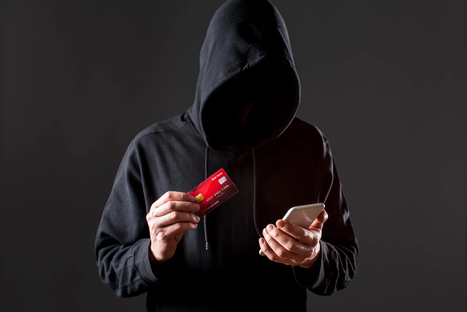 Persoană cu glugă ținând un telefon și un card bancar, simbolizând frauda.