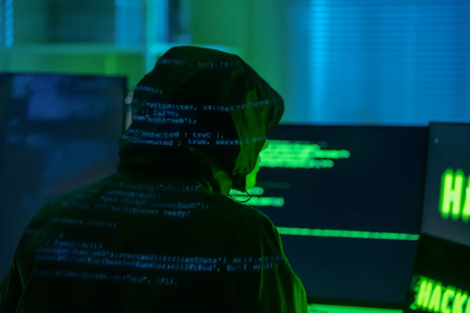 Un hacker lucrează la un computer, înconjurat de ecrane cu coduri verzi.