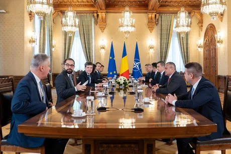 Discuții politice la o masă lungă, cu lideri și steaguri naționale.