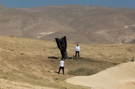 Două persoane în fața unui peisaj arid, interacționând cu un obiect metalic.