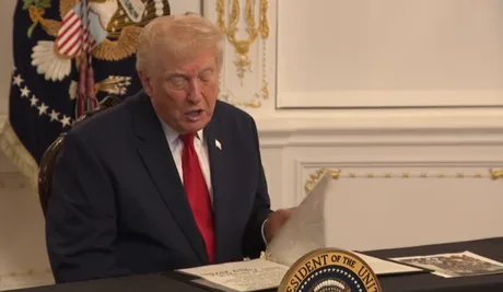 Donald Trump citind un document într-un cadru oficial, cu steagul în fundal.