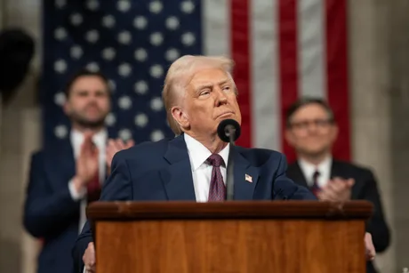Donald Trump la o conferință, cu steagul SUA în fundal.