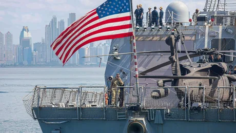 Nava militară americană în port, cu steagul SUA fluturând.