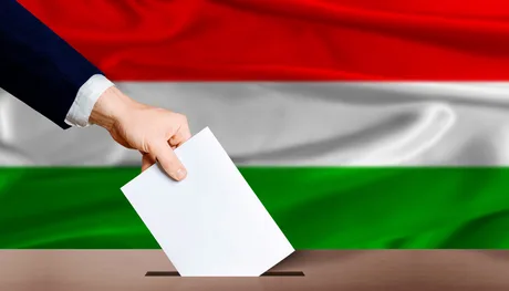O mână introduce un buletin de vot într-o urnă, cu steagul Ungariei în fundal.
