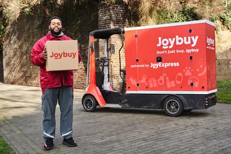 Un livrator zâmbitor cu o cutie Joybuy lângă un vehicul de livrare.