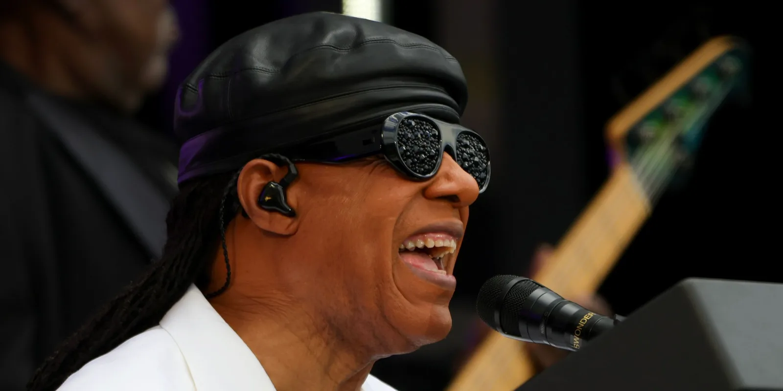 Stevie Wonder cântând pe scenă, purtând ochelari de soare și pălărie.