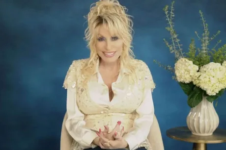 Dolly Parton zâmbind, îmbrăcată elegant, cu flori albe în fundal.