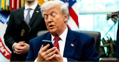 Donald Trump consultând un telefon în biroul oval, cu steagul american în fundal.