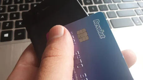 Card Revolut ținut în mână, pe fundal un laptop.