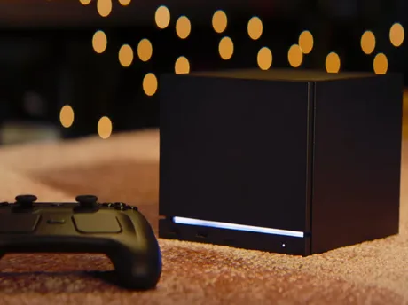 Dispozitiv de gaming negru cu un controller pe un covor, în fundal lumini difuze.