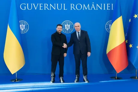 Zelenski și Bolojan strâng mâna în fața drapelului României.