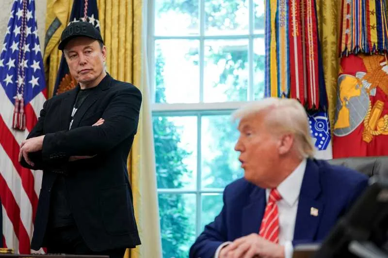 Elon Musk și Donald Trump discutând în Biroul Oval, cu steaguri în fundal.