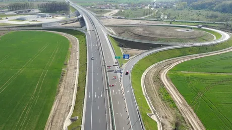 Vedere aeriană a unei autostrăzi în construcție, cu câmpuri verzi în jur.