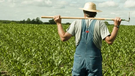 Un agricultor în câmp, pregătit pentru muncă, cu un zâmbet pe față.
