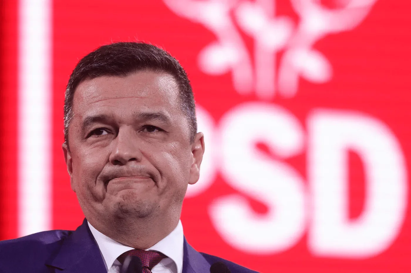 Sorin Grindeanu cu o expresie serioasă, în fața unui fundal roșu.