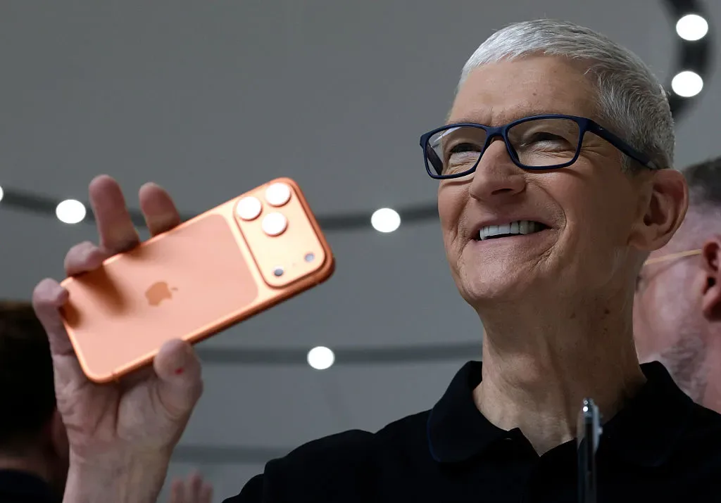 Un bărbat zâmbitor ține un telefon Apple portocaliu în mână, într-un mediu modern.