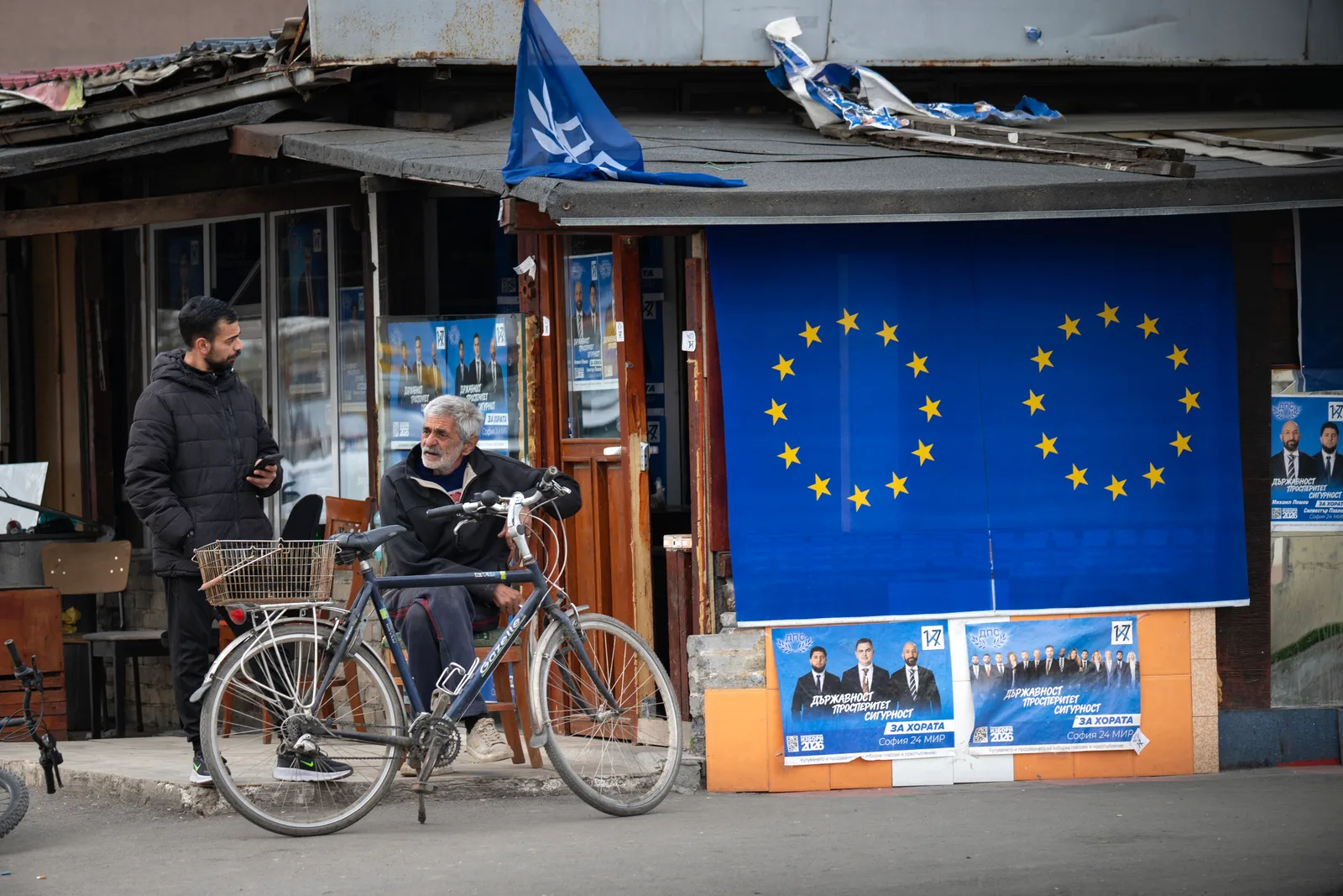 Oameni discutând lângă un magazin cu steagul Uniunii Europene expus.