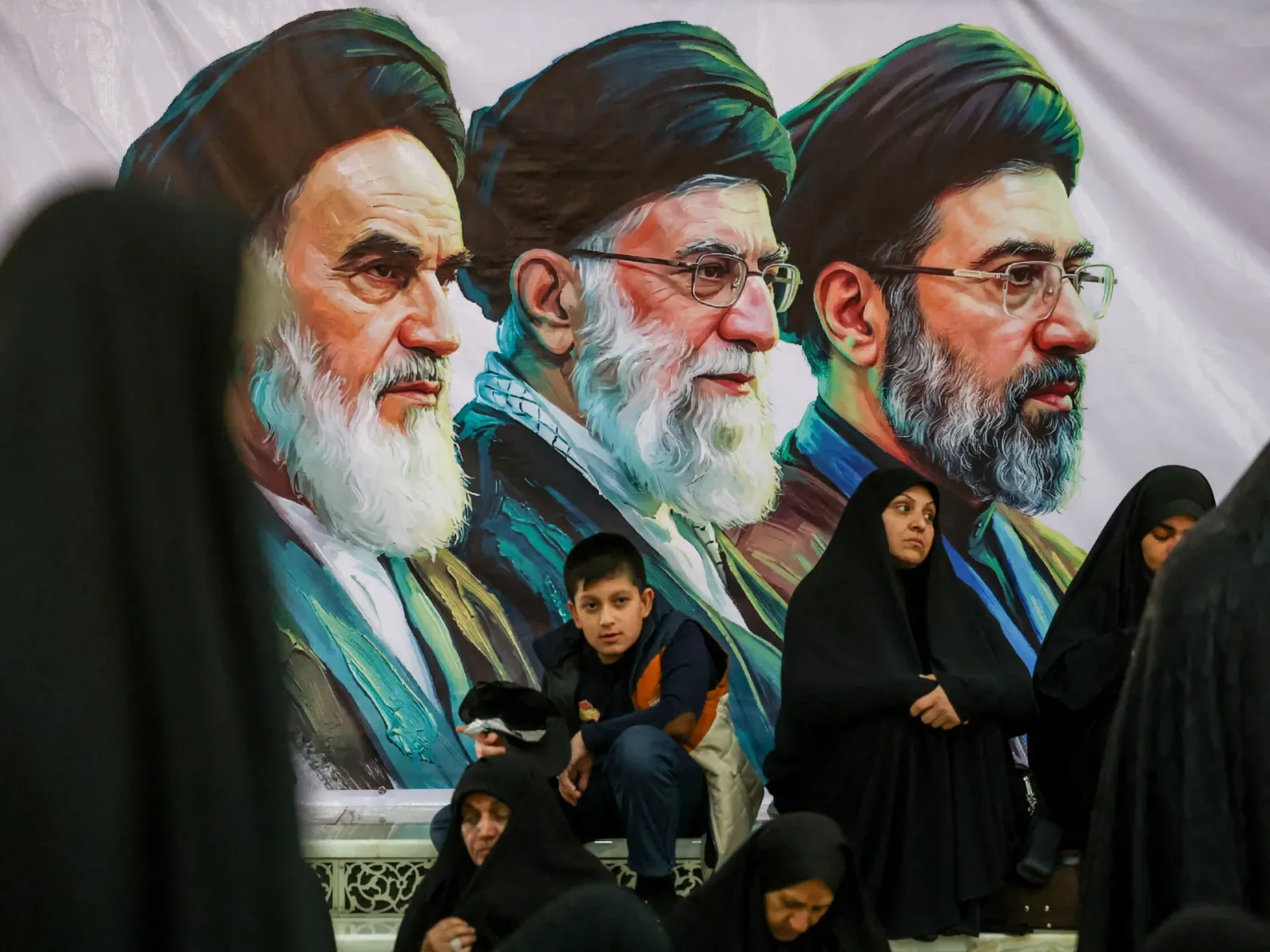 Oameni ascultând discursuri în fața unui mural cu lideri iranieni.